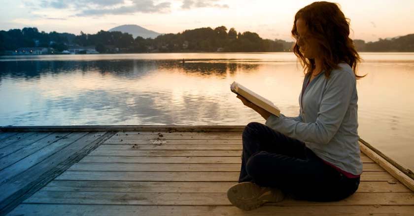 mujer sentada en un muelle orilla de un lago leyendo la biblia en mano ocaso