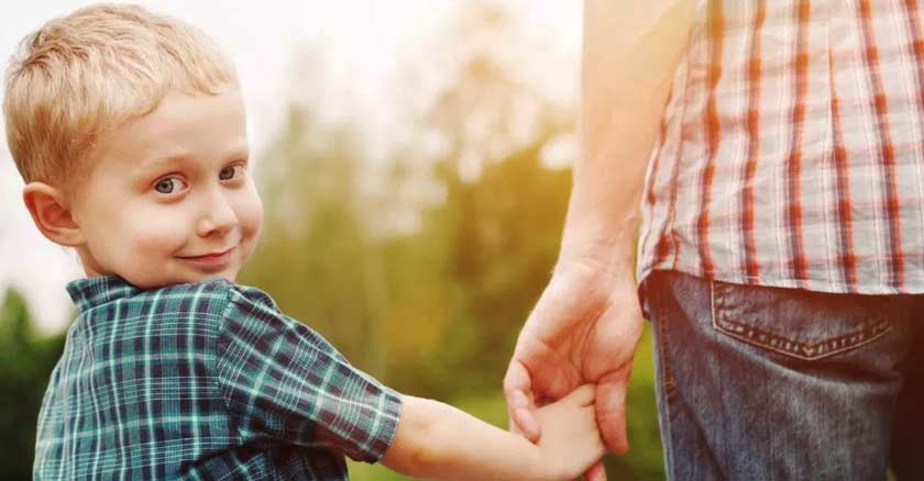 hijo sonriendo tomado de la mano de su papa mirando hacia la camara