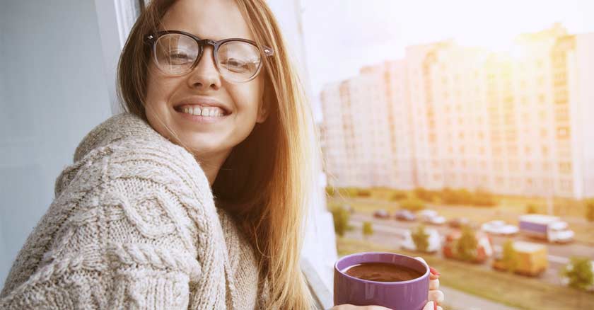 mujer sonriendo feliz mirando por la ventana de su casa apartamento ciudad taza en mano